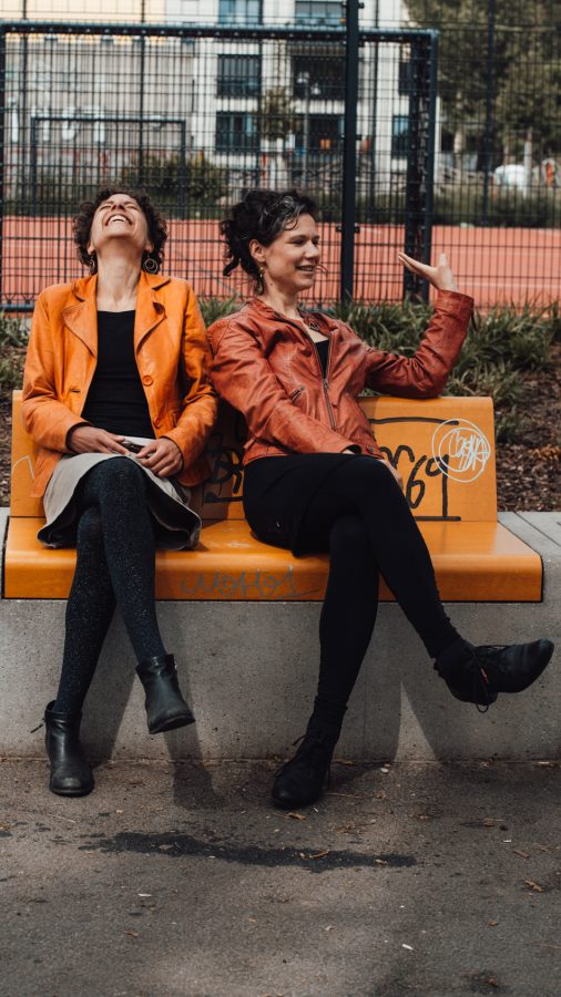 Duo Madame Unverfroren: zwei Musikerinnen in Lederjacken sitzen lachend auf einer orangefarbenen Bank
