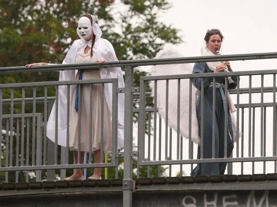 Zwei Darstellerinnen in weißen Kostümen, eine mit Maske, auf einer Brücke